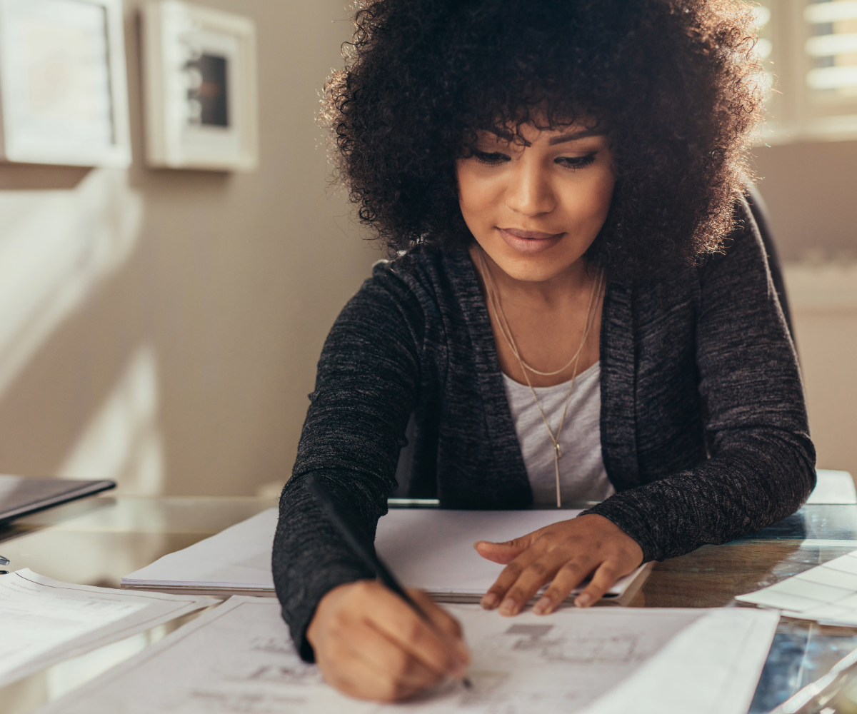 The image shows a woman with curly hair sitting at a desk, focused on drawing or writing on sheets of paper. She is wearing a dark gray cardigan and a light-colored blouse. Her left hand holds the paper steady while her right hand moves a pen across the paper. The desk is made of glass, and several other sheets of paper are visible on it. In the background, there are framed pictures on the wall and soft, natural light entering through window blinds, creating gentle shadows on the wall.